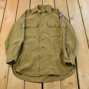 Può includere: Camicia verde oliva in stile militare con due tasche sul petto. Presenta toppe sulle maniche e sulle spalle, tra cui gradi e un emblema circolare. La camicia è stesa su una superficie di legno.