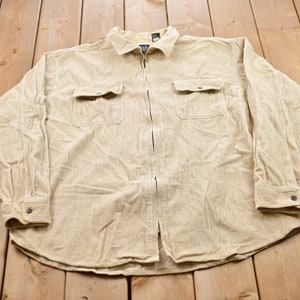 May include: A beige corduroy shirt with a zip fastening and two flap pockets. The shirt has long sleeves and a collared neckline.