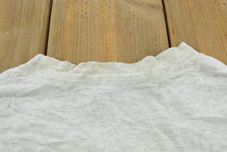 Op de afbeelding: Een lichtgrijs gem&ecirc;leerd t-shirt met een geribbelde ronde halslijn met zichtbare rafels en slijtage langs de rand van de kraag. De stof heeft een gespikkelde textuur, kenmerkend voor een klassiek gem&ecirc;leerd katoenmengsel.
