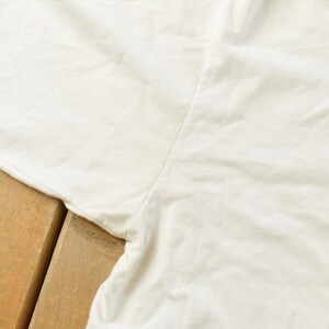 May include: Close-up of a white garment, possibly a t-shirt or top, with visible seams and fabric texture. The garment is slightly wrinkled, suggesting it is made of a soft material. The background is a wooden surface.