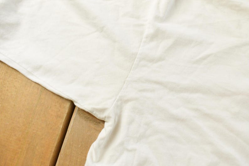 May include: Close-up of a white garment, possibly a t-shirt or top, with visible seams and fabric texture. The garment is slightly wrinkled, suggesting it is made of a soft material. The background is a wooden surface.