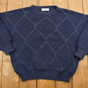 May include: A navy blue knitted sweater with a diamond pattern. The sweater has a crew neck and long sleeves.