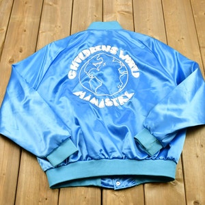 May include: Light blue satin jacket with a snap closure and light blue ribbed cuffs and collar. The back of the jacket features the white text "CHILDRENS WORLD MINISTRY" surrounding a globe graphic.