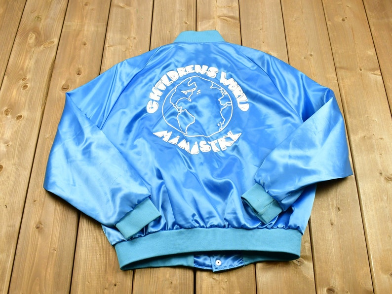 May include: Light blue satin jacket with a snap closure and light blue ribbed cuffs and collar. The back of the jacket features the white text "CHILDRENS WORLD MINISTRY" surrounding a globe graphic.