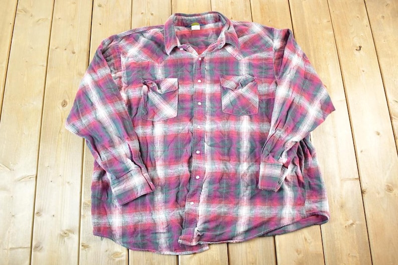 Puede incluir: Una camisa de cuadros de manga larga en tonos rojos, blancos y turquesas. La camisa tiene dos bolsillos en el pecho y una botonadura frontal. La tela tiene un aspecto suave y desgastado, lo que sugiere que es vintage.