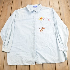 Può includere: Una camicia a maniche lunghe di colore azzurro chiaro con bottoni e ricami di Winnie the Pooh e Tigger. La camicia ha una tasca sul lato sinistro con un disegno ricamato in rosso.