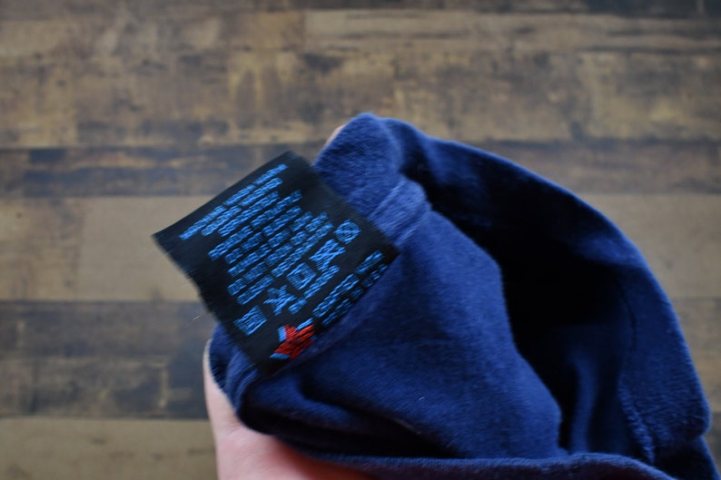 May include: Close-up of a navy blue t-shirt with a black care label. The label has text in English, French, German, Spanish, Italian, and Japanese. The label also has symbols for washing, drying, and ironing.