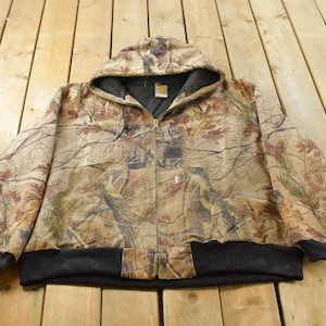 Peut inclure: Une veste à capuche zippée Carhartt avec un motif camouflage marron, beige et vert. La veste présente un ourlet et des poignets côtelés noirs, une doublure intérieure noire et une petite étiquette de marque blanche sur la poche avant.