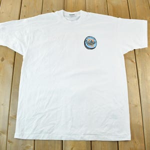 May include: White t-shirt with a crew neck and short sleeves. A circular graphic on the left chest features the word "Vintage" in a stylized font, surrounded by a blue and yellow design. The shirt is laid flat on a wooden surface.
