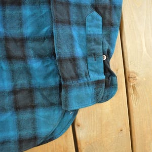May include: Close-up of a blue and black plaid flannel shirt. The shirt features a buttoned cuff and a buttoned sleeve placket. The fabric has a soft, textured appearance.