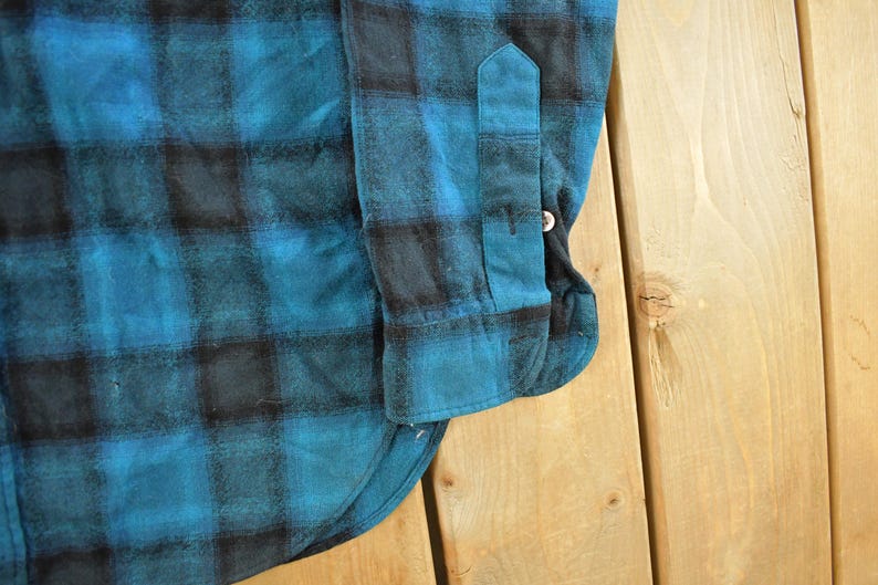 May include: Close-up of a blue and black plaid flannel shirt. The shirt features a buttoned cuff and a buttoned sleeve placket. The fabric has a soft, textured appearance.