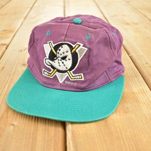 Può includere: Cappellino da baseball viola e verde acqua con il logo degli Anaheim Ducks. Il cappellino ha una visiera verde acqua e una corona viola. Il logo presenta una maschera da hockey e bastoni da hockey incrociati, un accessorio sportivo.