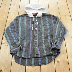 May include: A grey button-down shirt with vertical teal and purple stripes. It features a grey hood with white drawstrings. The shirt is made of a soft, textured fabric. The shirt is a casual style.