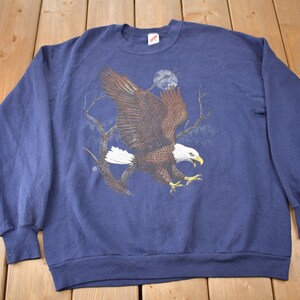 K&ouml;nnte beinhalten: Ein marineblauer Sweatshirt mit einem Aufdruck eines Wei&szlig;kopfseeadlers im Flug vor einem mondbeschienenen Himmel.