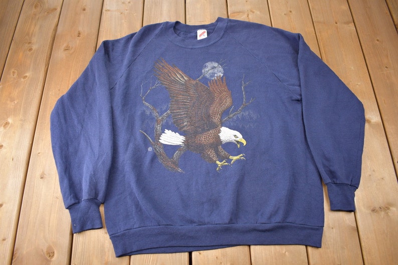 K&ouml;nnte beinhalten: Ein marineblauer Sweatshirt mit einem Aufdruck eines Wei&szlig;kopfseeadlers im Flug vor einem mondbeschienenen Himmel.