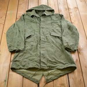 Puede incluir: Chaqueta de camuflaje verde con capucha y dos bolsillos delanteros. La chaqueta tiene un cordón en la cintura y cierre de botones. La tela tiene un pequeño patrón cuadrado con manchas verdes oscuras.