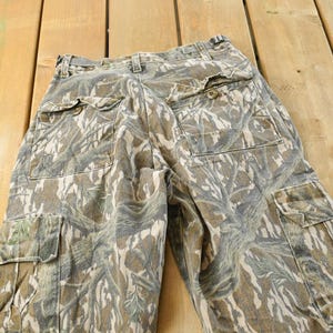 May include: Camouflage cargo pants in shades of green, brown, and tan. The pants feature multiple pockets, a button and zipper closure, and a belt loop waistband. The fabric has a textured appearance.