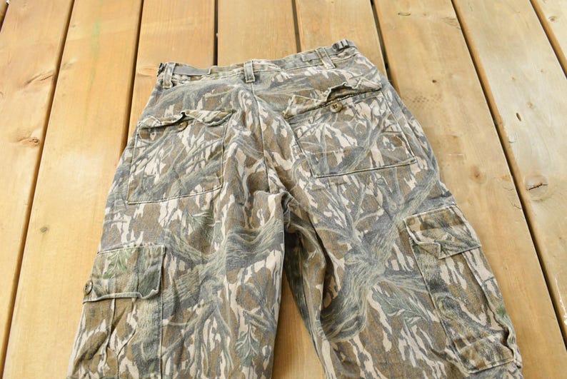 May include: Camouflage cargo pants in shades of green, brown, and tan. The pants feature multiple pockets, a button and zipper closure, and a belt loop waistband. The fabric has a textured appearance.