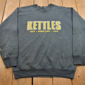 May include: A gray sweatshirt with a yellow graphic that says "KETTLES NORTH * BOURBON STREET * SOUTH".