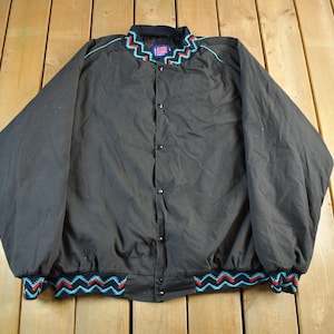 May include: Black snap-front jacket with a colourful zigzag pattern on the collar, cuffs, and hem. The jacket has a classic bomber style with a black collar and cuffs. The zigzag pattern includes red, turquoise, and white accents.