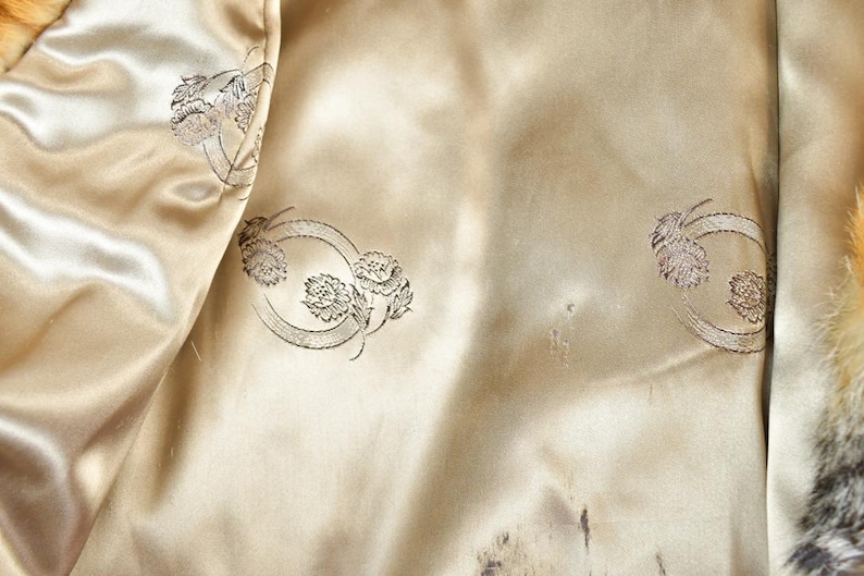 May include: Close-up of the interior lining of a vintage coat. The lining is a light beige satin with a subtle sheen. Decorative floral embroidery in a circular pattern is visible. The coat's fur trim is visible on the right side.