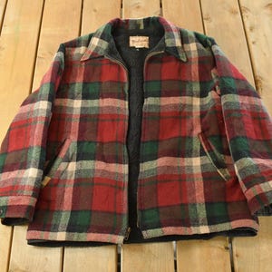 May include: A vintage plaid jacket in red, green, and cream. The jacket features a zip closure, two front pockets, and a dark grey lining. The jacket is made of a thick, warm material, suitable for cooler weather.