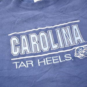 May include: A blue t-shirt with the text "CAROLINA TAR HEELS" in white and light blue lettering. A blue and white mascot is in the bottom right corner.