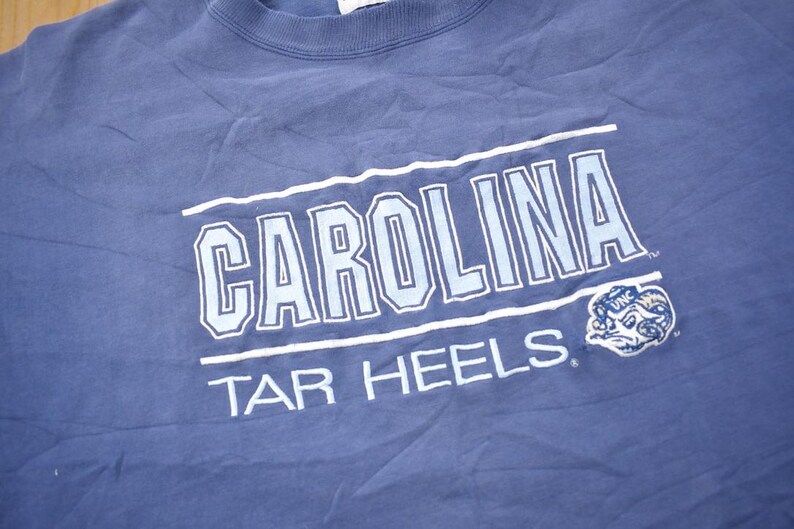 May include: A blue t-shirt with the text "CAROLINA TAR HEELS" in white and light blue lettering. A blue and white mascot is in the bottom right corner.
