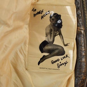 May include: Close-up of the inside of a vintage brown leather jacket. A cream-colored lining features a printed pin-up girl graphic with the text "To Avirex Aces" and "Good Luck, Jimy".