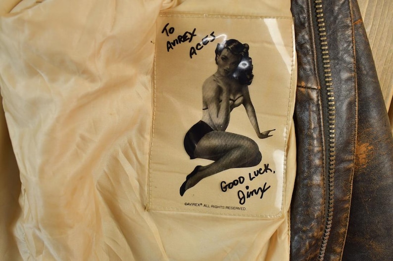 May include: Close-up of the inside of a vintage brown leather jacket. A cream-colored lining features a printed pin-up girl graphic with the text "To Avirex Aces" and "Good Luck, Jimy".