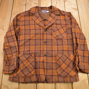 Könnte beinhalten: Eine braune und orangefarbene karierte Jacke mit Knopfleiste und zwei großen Taschen. Die Jacke hat einen Kragen und lange Ärmel.