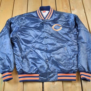 Op de afbeelding: Een blauwe satijnen bomberjack met oranje en witte strepen aan de manchetten en tailleband. De jas heeft een drukknopsluiting en een groot "C"-logo op de linkerborst.