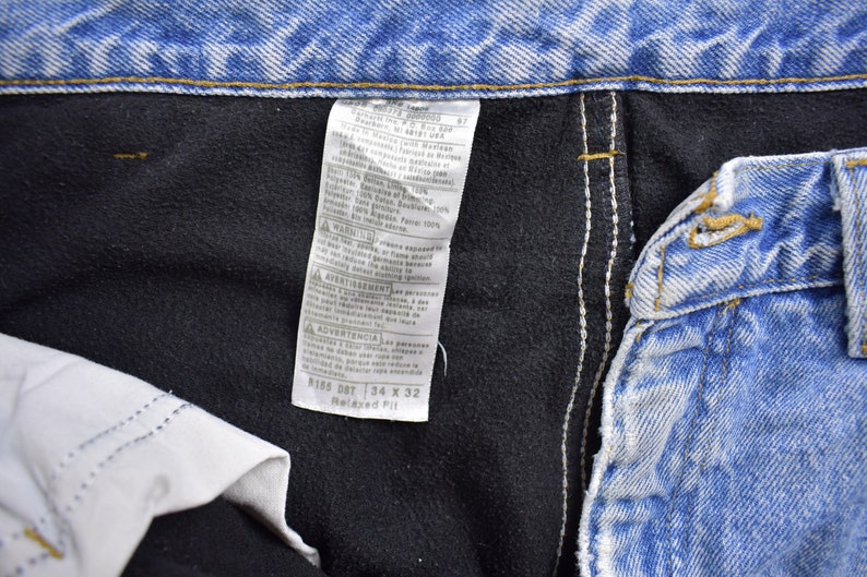 May include: A close-up of a blue denim jean label with white stitching. The label has text that reads "8165 08T 34 X 32 Relaxed Fit".