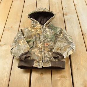 May include: A hooded jacket featuring a camouflage pattern in shades of brown, green, and tan. The jacket has a dark brown ribbed hem and sleeve cuffs, a dark brown interior lining, a front zipper closure, and a small rectangular Carhartt brand patch.