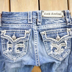 K&ouml;nnte beinhalten: Eine blaue Jeanshose mit wei&szlig;en N&auml;hten und Verzierungen. Die Ges&auml;&szlig;taschen haben ein abgewetztes Design mit einer silbernen Fleur-de-lis und Strasssteinen. Die Jeans hat ein "Rock Revival"-Etikett am hinteren Bund.