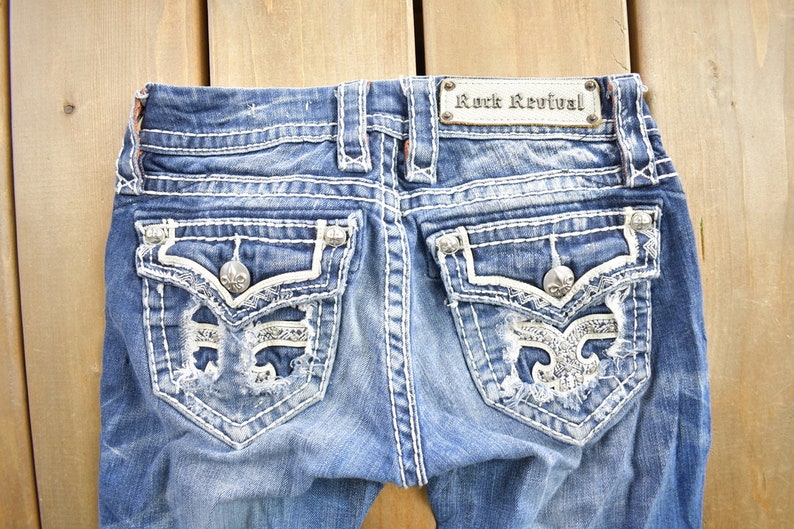 K&ouml;nnte beinhalten: Eine blaue Jeanshose mit wei&szlig;en N&auml;hten und Verzierungen. Die Ges&auml;&szlig;taschen haben ein abgewetztes Design mit einer silbernen Fleur-de-lis und Strasssteinen. Die Jeans hat ein "Rock Revival"-Etikett am hinteren Bund.