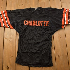 Op de afbeelding: Zwart mesh jersey met oranje en witte strepen op de mouwen en het woord "CHARLOTTE" in oranje op de voorkant.