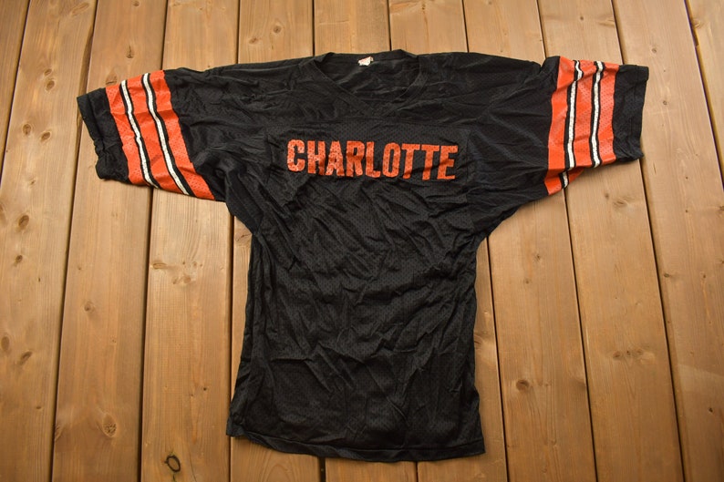 Op de afbeelding: Zwart mesh jersey met oranje en witte strepen op de mouwen en het woord "CHARLOTTE" in oranje op de voorkant.