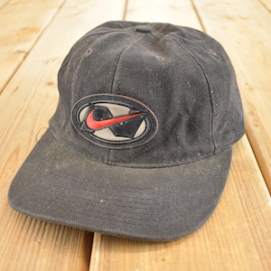 Op de afbeelding: Zwarte baseballcap met een gebogen klep. De cap heeft een Nike-logo in een witte ovaal met een rode swoosh en zwarte details. De cap is gemaakt van een versleten, getextureerde stof.