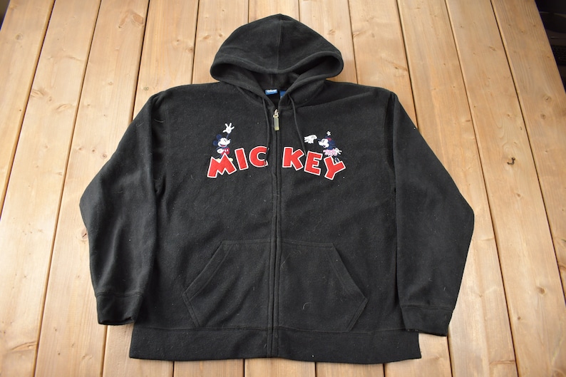 May include: Black fleece hoodie with a zipper closure and a Mickey Mouse graphic on the front. The graphic features Mickey Mouse and his friends in a line, with the word "Mickey" in red letters and the word "Key" in black letters.
