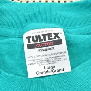 May include: A teal t-shirt featuring a white interior neck label with the brand name TULTEX in black text. The label specifies 100% cotton, preshrunk, size Large, and indicates the fabric was made in the U.S.A. and sewn in Mexico for the garment.