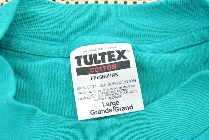 May include: A teal t-shirt featuring a white interior neck label with the brand name TULTEX in black text. The label specifies 100% cotton, preshrunk, size Large, and indicates the fabric was made in the U.S.A. and sewn in Mexico for the garment.