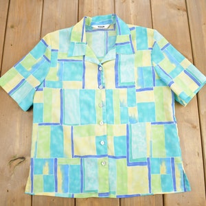 Può includere: Una camicia a maniche corte con bottoni e un motivo geometrico in tonalità di blu, verde e giallo. La camicia ha un colletto e una chiusura con bottoni.
