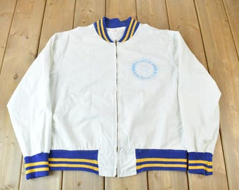May include: White varsity jacket with blue and yellow striped trim. The jacket has a zipper closure and a blue embroidered logo on the front.