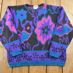 Puede incluir: Un suéter de punto con estampado floral morado, rosa y turquesa sobre fondo negro. El suéter tiene cuello redondo y mangas largas.