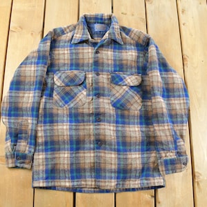 Op de afbeelding: Een vintage geruit flanellen overhemd in blauw, bruin en beige. Het shirt met lange mouwen heeft twee borstzakken en een kraag. Het shirt ligt plat op een houten oppervlak.