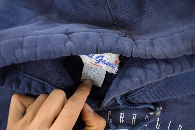 K&ouml;nnte beinhalten: Nahaufnahme eines marineblauen Kapuzenpullovers. Das Etikett lautet "The Game" und "X-Large". Der Pullover hat einen Kordelzug und das Wort "CAROLINA" ist in wei&szlig;en Buchstaben zu sehen.