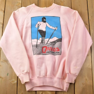 May include: Pink sweatshirt featuring a graphic of a person skiing in Davos, Switzerland. The graphic is in black, white and blue.