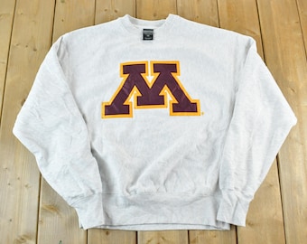 Könnte beinhalten: Hellgraues Sweatshirt mit großem weinrotem und goldenem "M"-Logo auf der Vorderseite. Das Sweatshirt hat einen Rundhalsausschnitt und gerippte Bündchen und Saum. Das Logo ist ein kühnes Design im College-Stil.