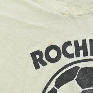 May include: A cream-colored t-shirt featuring a black graphic print of a soccer ball and the partial text ROCHEST in a bold, sans-serif font. The garment has a ribbed crew neckline and a soft, lightweight fabric construction.